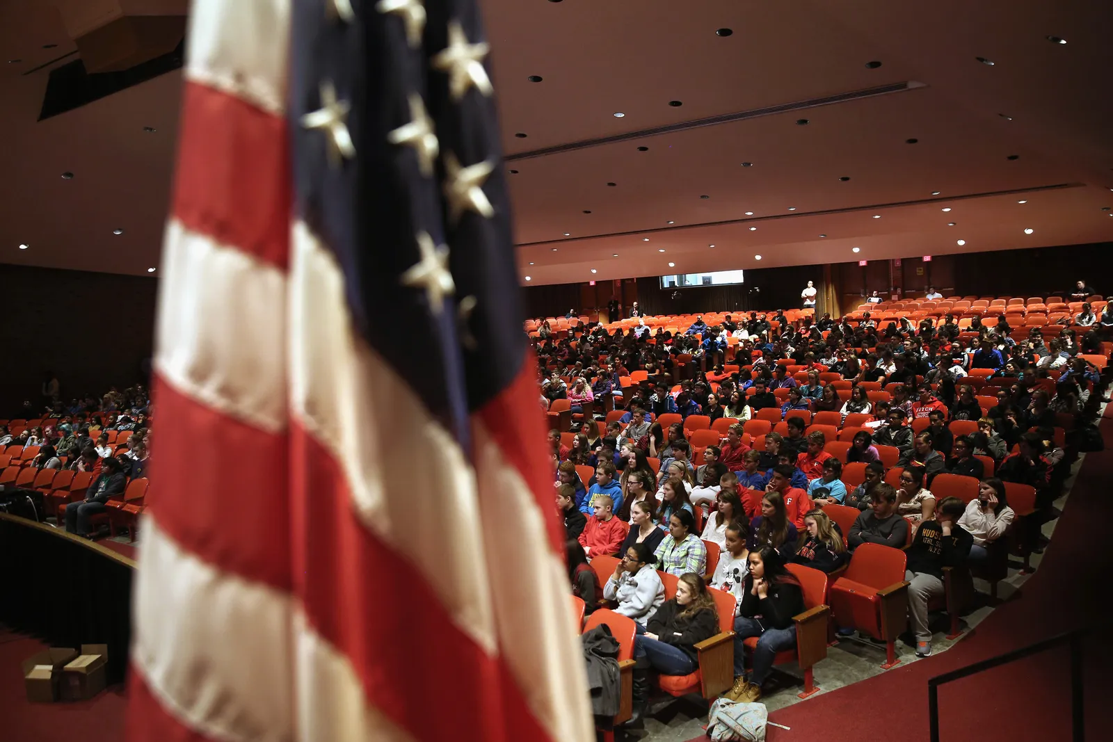 An auditorium is filled with high school students framed by a US flag in the foreground.