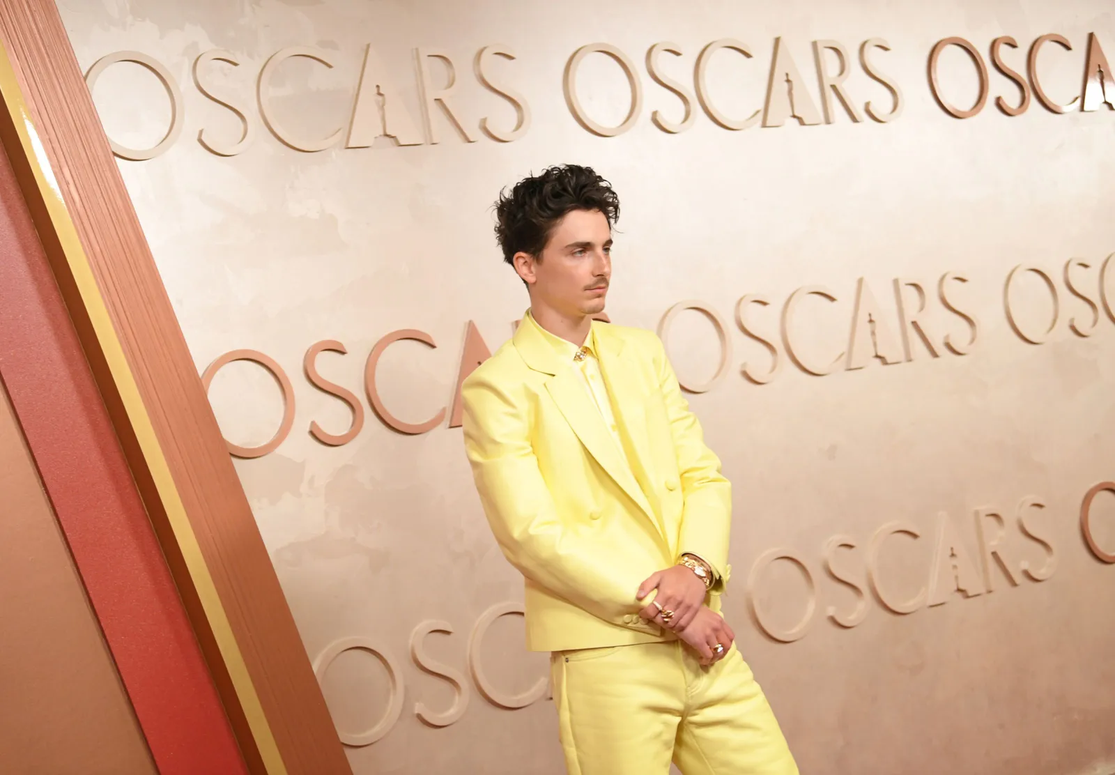 A man in a monochrome canary yellow suit crossed his hands in front of him while standing in front of a sign that says Oscars.