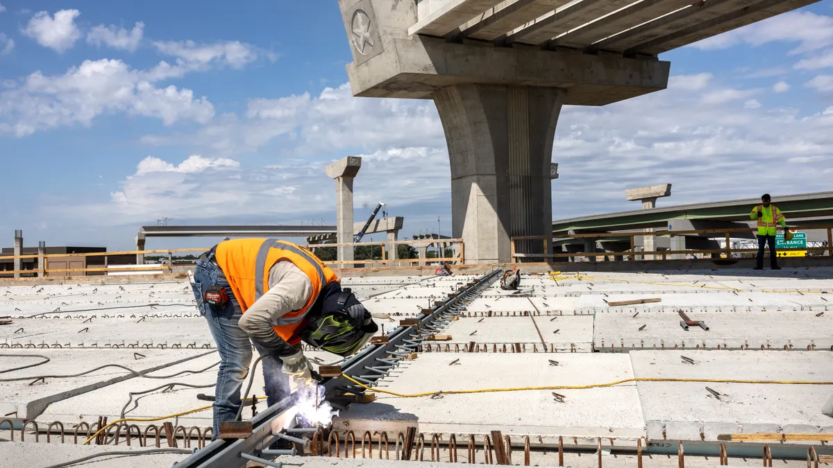 A person in a reflective vest and utility mask bends over their work, with unfinished concrete bridge parts in the background.