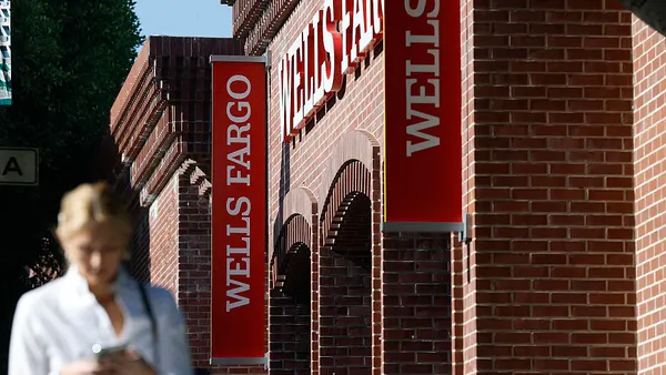 A person on their phone walks past a brick Wells Fargo branch with red signage.