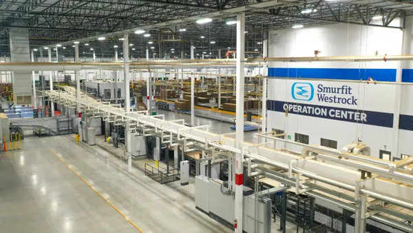 Industrial equipment and boxes are seen from an aerial view inside a facility that says Smurfit Westrock operation center on the wall.