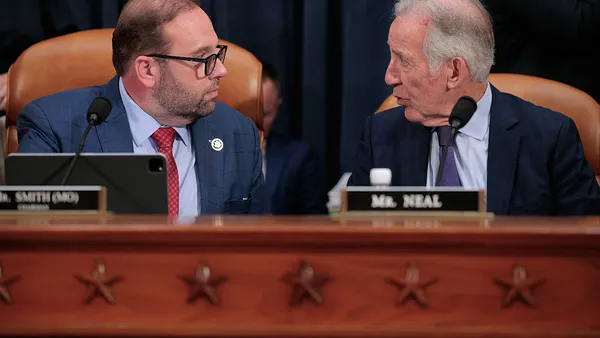 Two people are seated at a table with microphones in a room. They are looking at each other