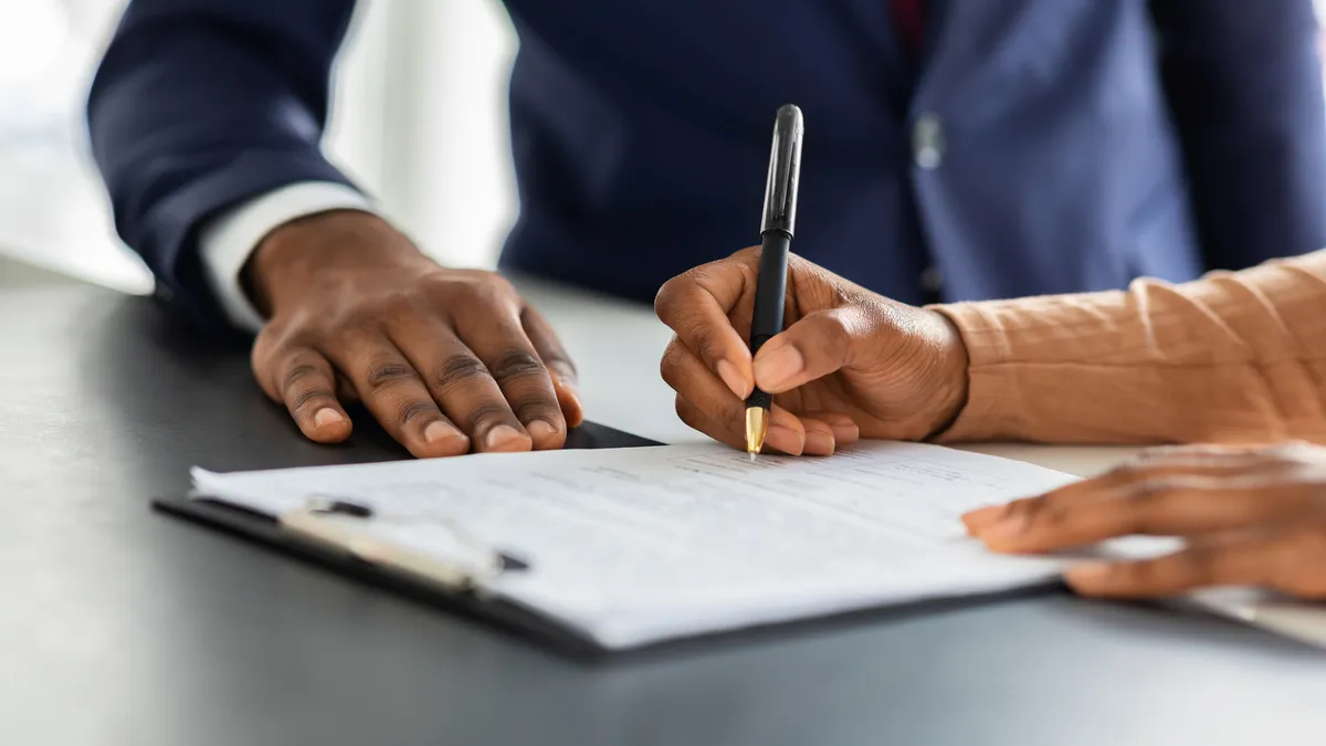 Two people sitting at a table, with only hands visible. One is writing on a pad of paper with a pen.