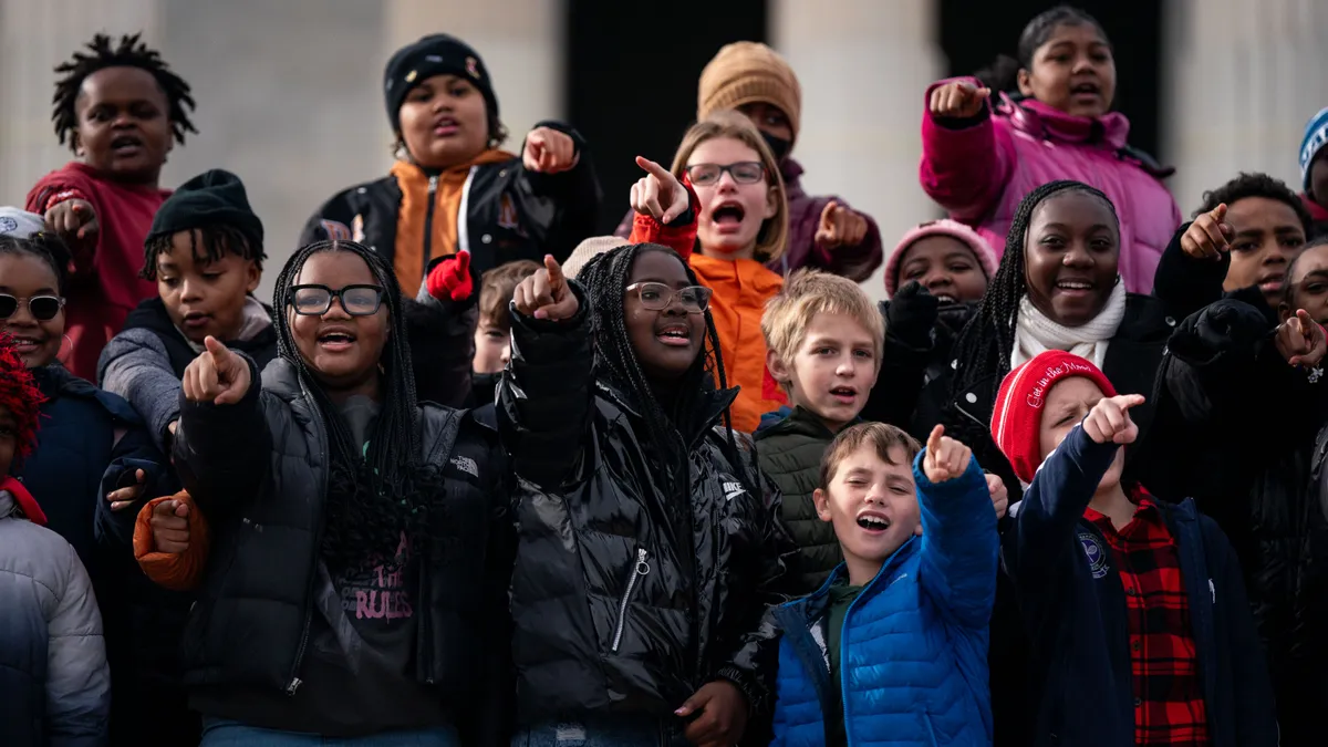 A group of students stand outside on different levels of steps outside. They are dressed in coats and some are pointing. Some have their mouths open as if they are talking or singing.