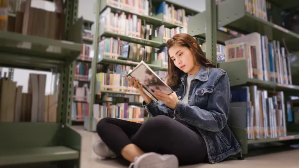 Student sits on floor in a library holding a book. Behind the student are rows of bookshelves.