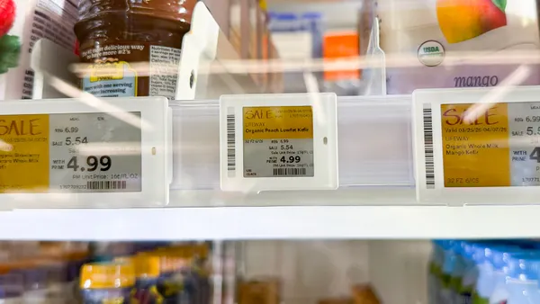 Close-up of electronic shelf labels in a refrigerated case displaying “SALE” prices for Lifeway kefir, including “Organic Peach Lowfat Kefir,” with pricing shown as “REG 6.99,” “SALE 5.54,” and “WITH PRIME 4.99,” and blurred dairy containers behind.
