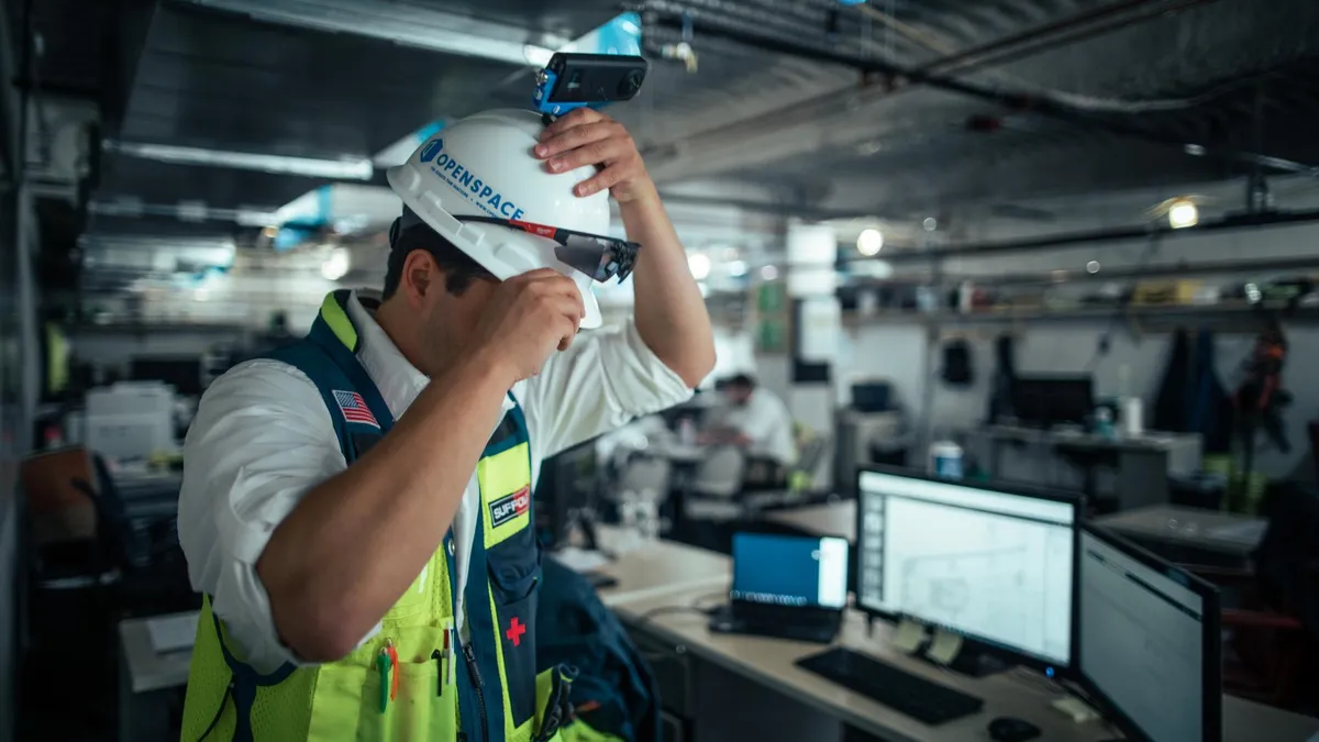 A man wears a yellow safety vest and a white hardhat with a camera mounted on the top. He sits in an industrial control room-esque room.