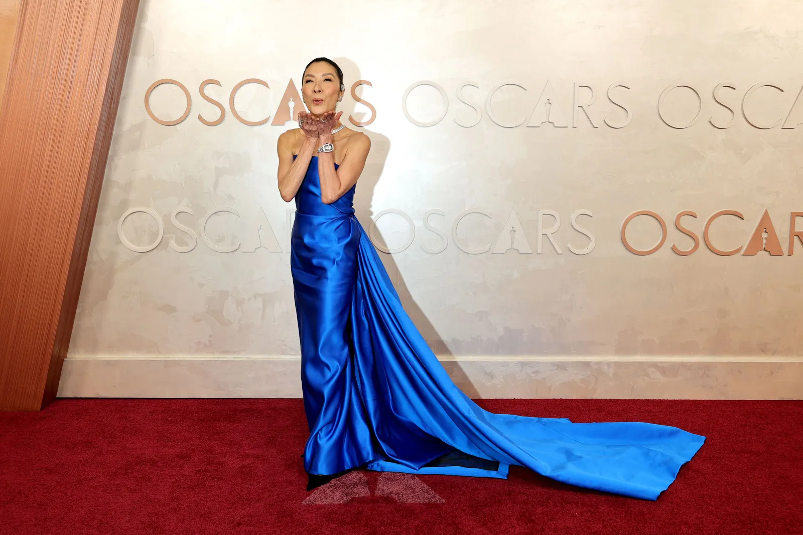 A woman in a strapless blue ballgown with a long train stands on the red carpet.