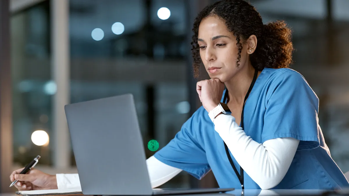 A healthcare worker looks at a laptop wile writing down information.