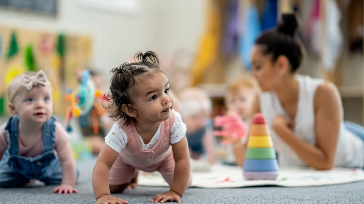 Two babies are in a crawl position on the floor looking to the side and ahead. In the background, an adult is on the floor with other children.