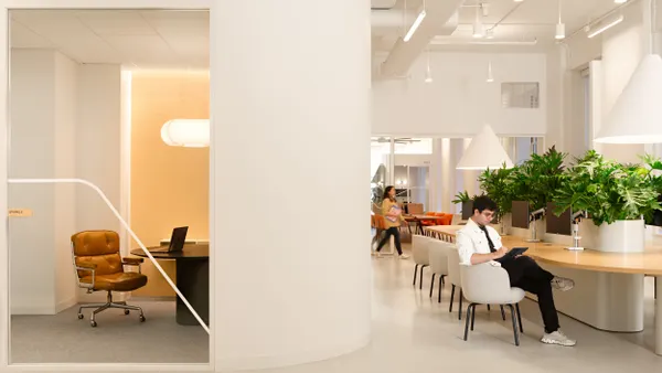 People work inside a white-walled office space with high ceilings and plants. One employee sits at a public table, while a small, more private space is seen to the left.