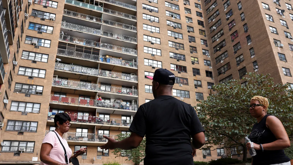 Three people speak with each other in front of a large multifamily building.