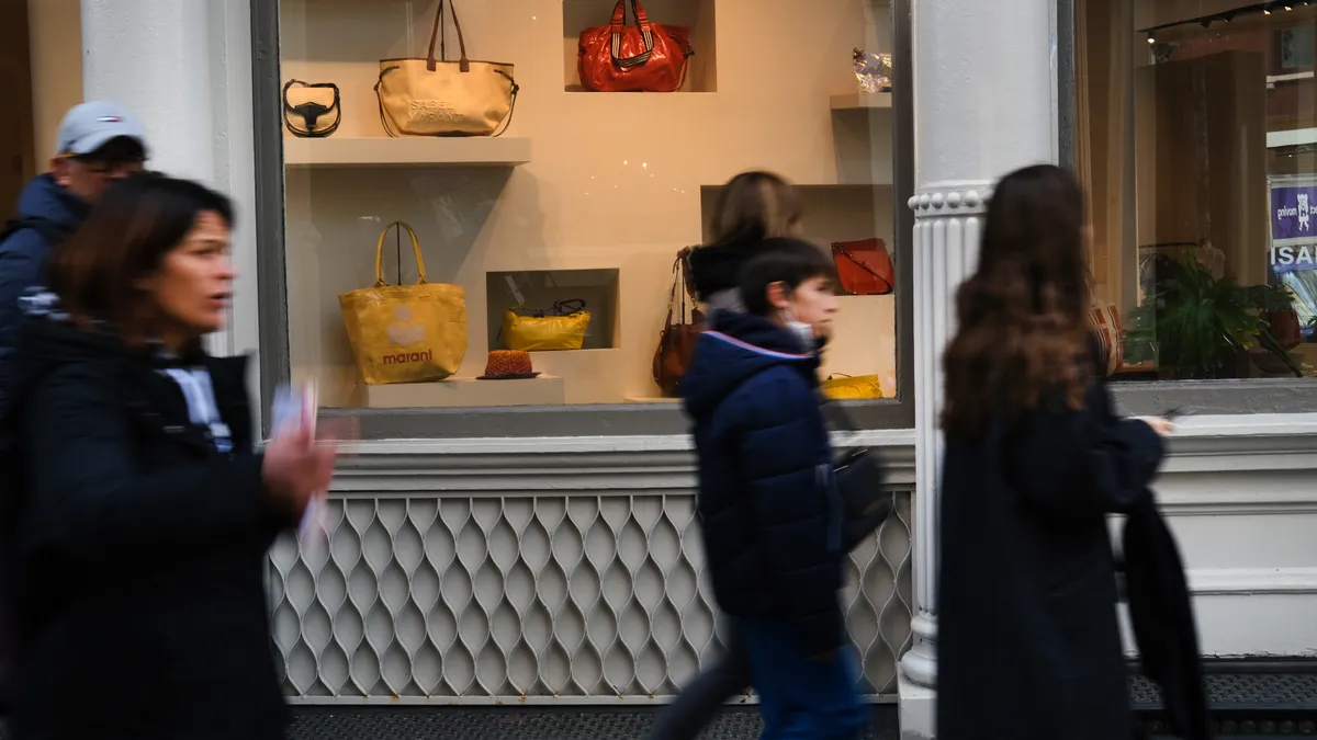 A group of people shopping walk quickly past a store window displaying luxury handbags.