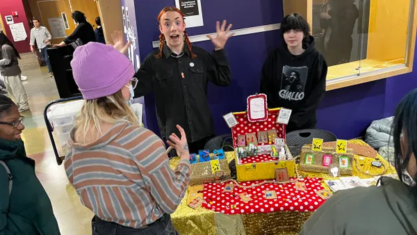 Students are gathered around a table in a school hallway. One student, facing the camera, has their hands up. There are colorful items on the table