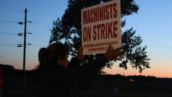 The back of a woman holding a red and white Machinists on strike sign at dusk.