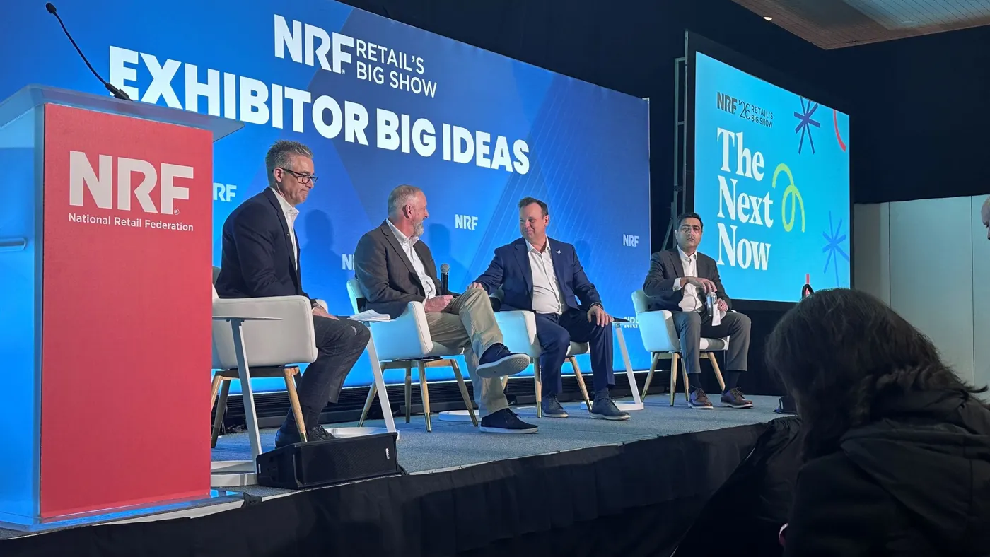 Panel with three men in suits on stage in front of a blue background that has a geometric pattern and reads NRF numerous times in small font.