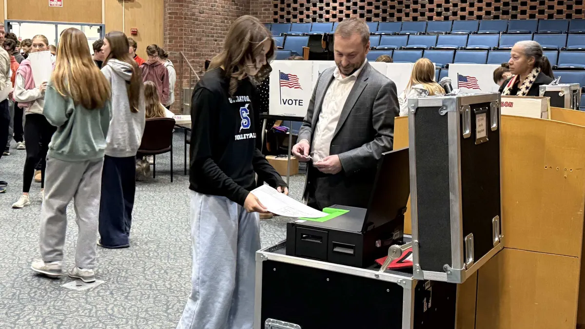 A student places a paper ballot into a voting machine as an adult stands next to the student.