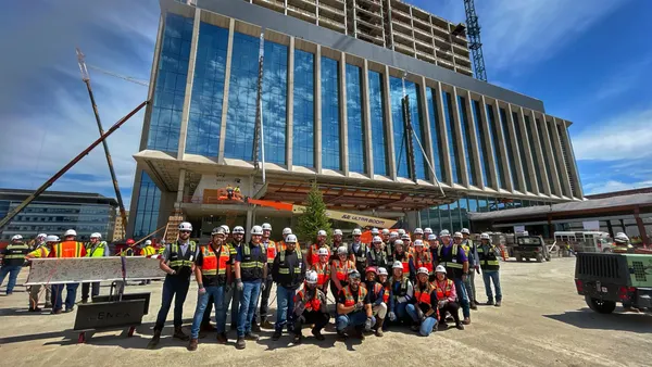 A crew of people in safety gear on a construction site pose in front of a building that's under construction, steel beams and girders visible.