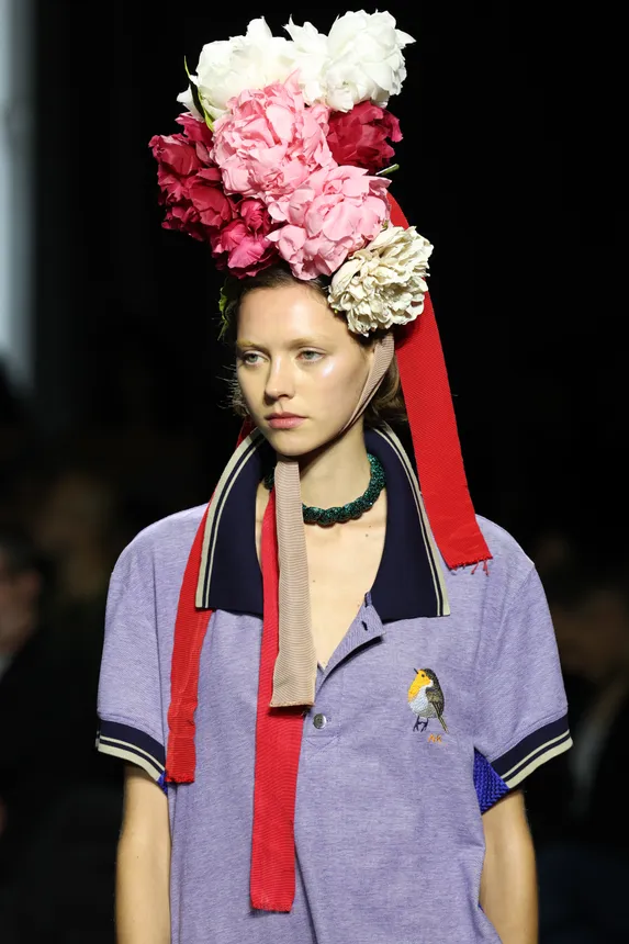 A model wears an elaborate headdress with flowers and a polo shirt.