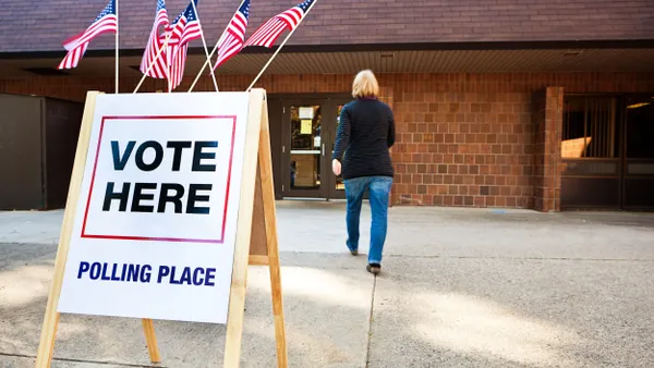 A sign with U.S. flags that says "Vote Here Polling Place" in the foreground with person walking into a building in the background.