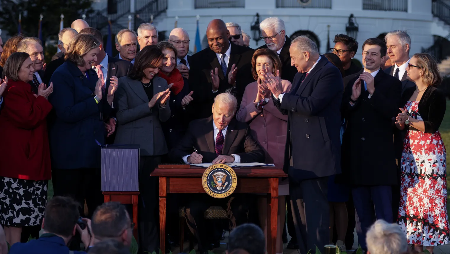 Man sitting at desk, signing a bill, surrounded by other people in front of the White House