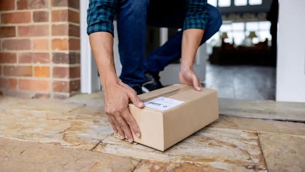 A person's hands are seen picking up a corrugated box that was left on a doorstep.