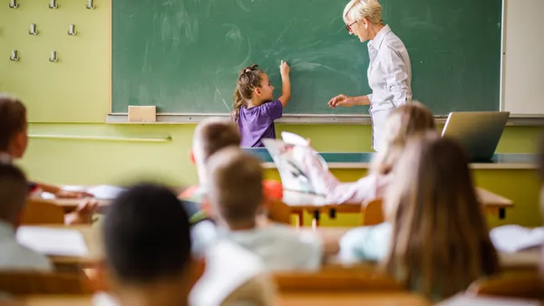 An adult and a young student stand in front of the classroom near a green chalkboard. Students seated at desk are facing them with their backs to the camera.