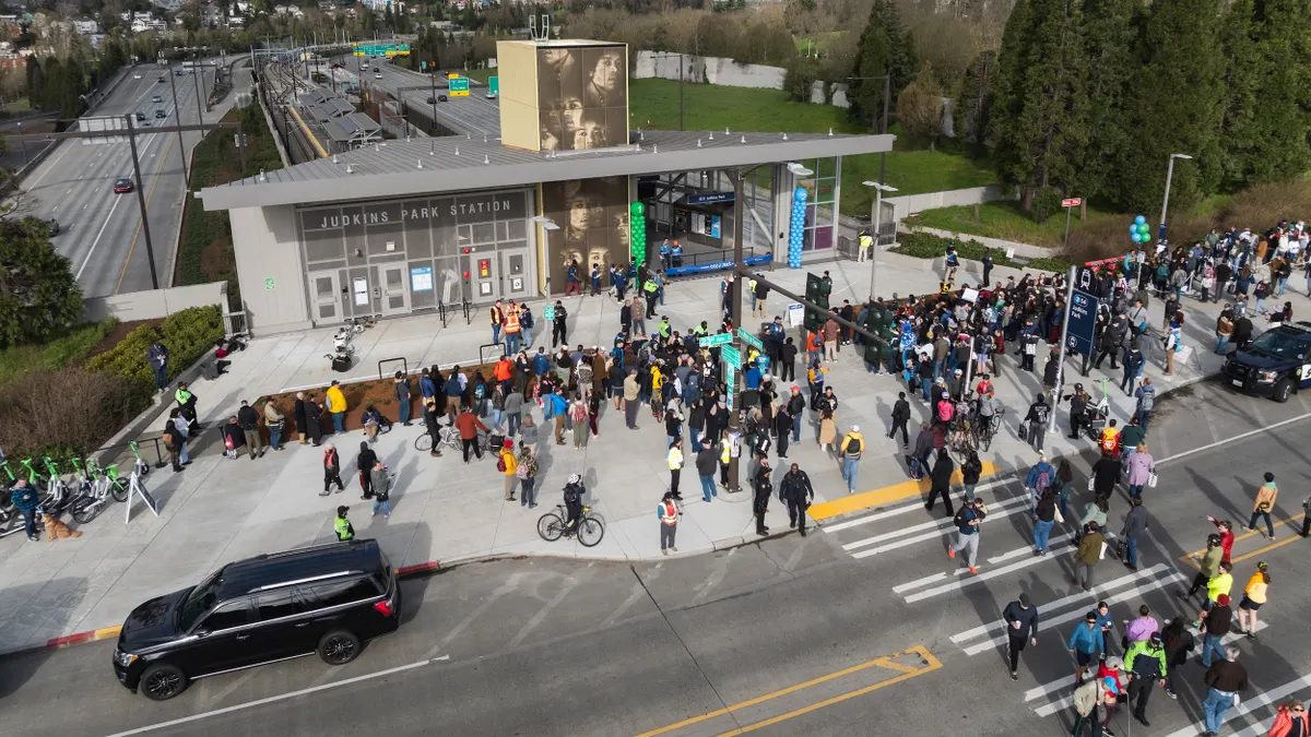 An aerial view of a train statin alongside a highway with crowds of people walking toward the gray station building.