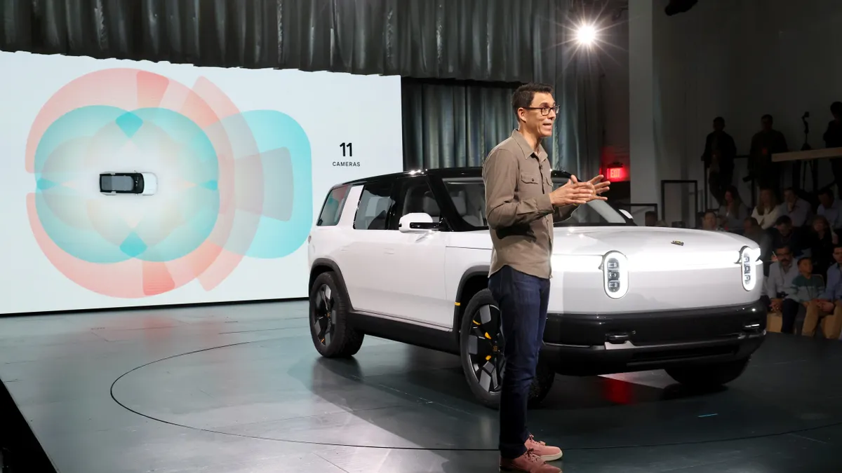 A man in glasses in front of a white car and a screen on stage.