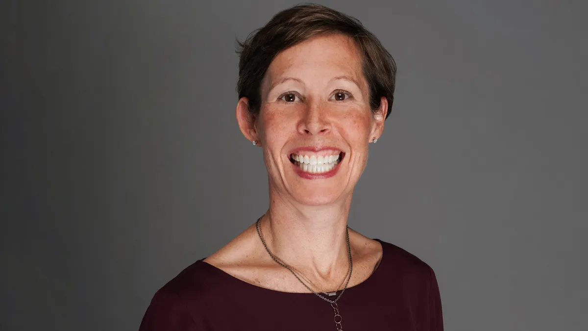 An image of a woman with short brown hair smiling in front of a gray background. She is wearing a red shirt.