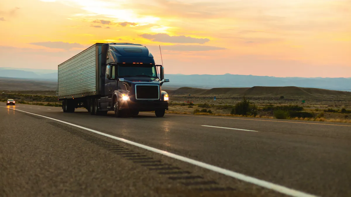 A tractor-trailer with headlights on in front of a car on an interestate with a desert backdrop.