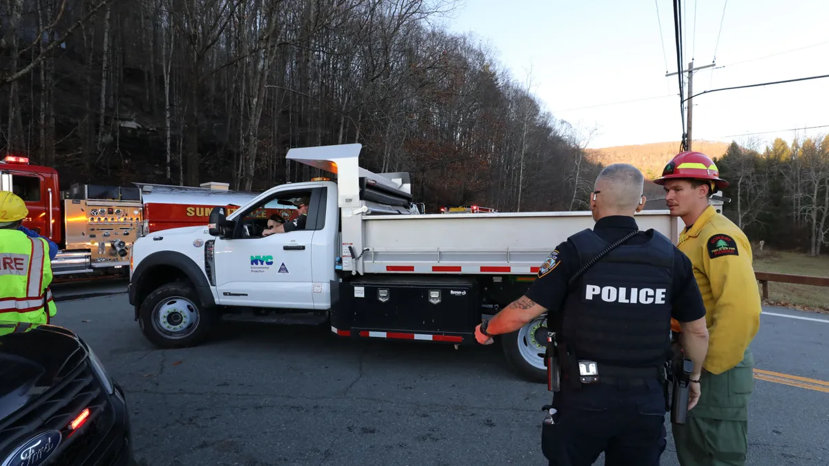 Two people in uniforms speak on a road. Several trucks including a fire truck are parked next to them. Behind the trucks are a forested area.