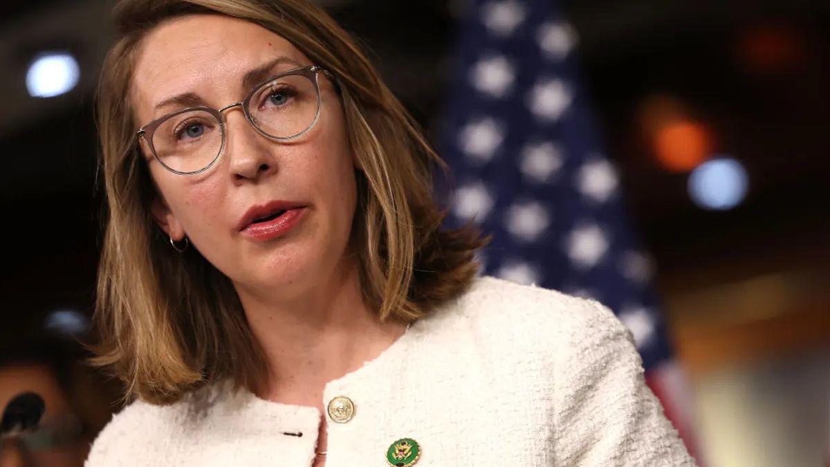 Hillary Scholten, a woman in a white jacket with glasses and shoulder-length hair, looks to the right standing in front of an American flag.