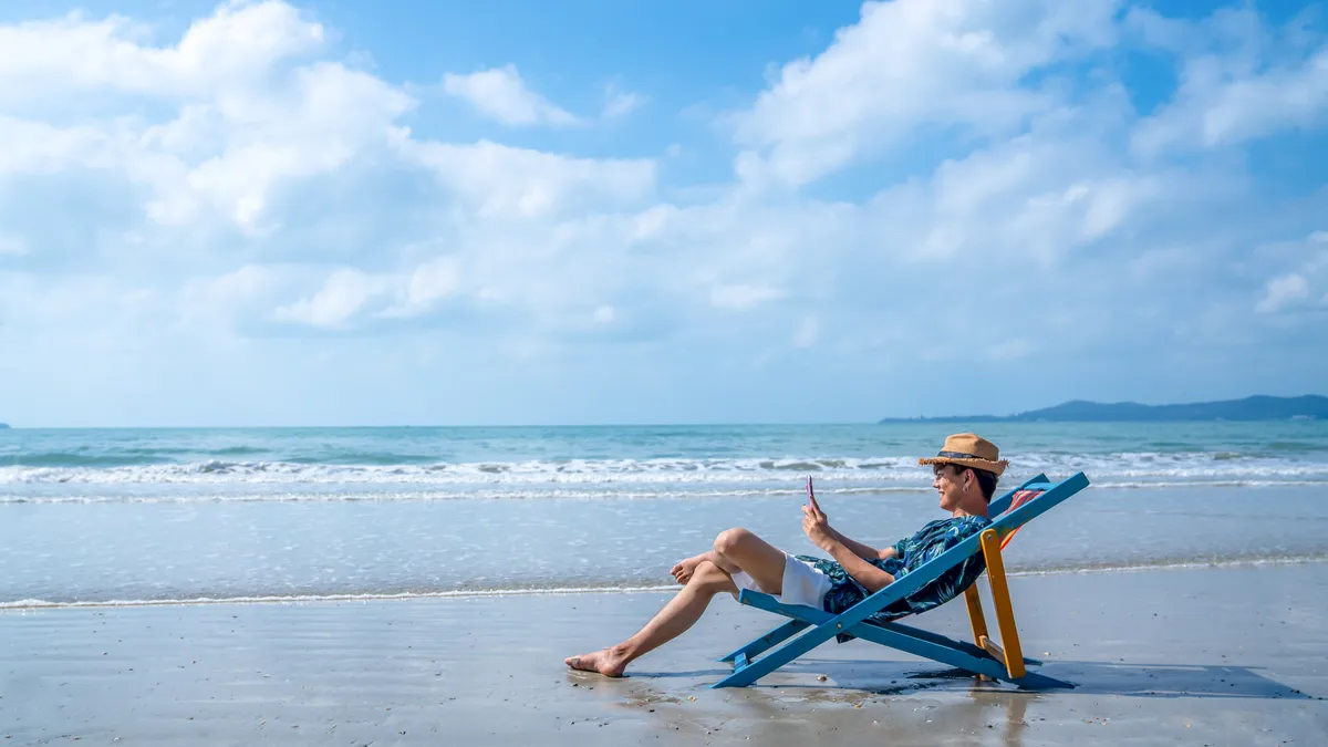 A person in vacation attire sits on a chair on a beach while using a smartphone to conduct a video call.