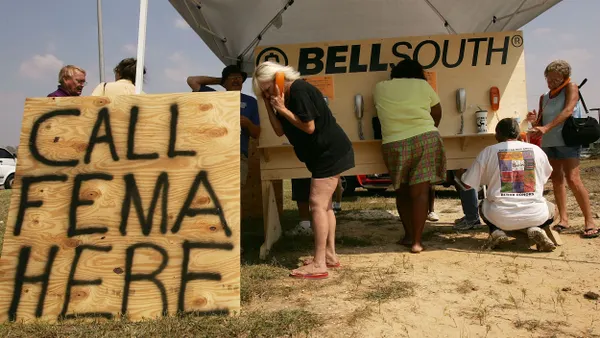 A person talks on a mobile phone in the foreground and others talk on phones in the background under a tent shelter with a wooden sign with painted message: "Call FEMA Here"