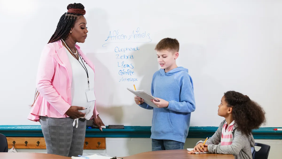 A pregnant teacher stands in front of the classroom whiteboard and speaks to a student