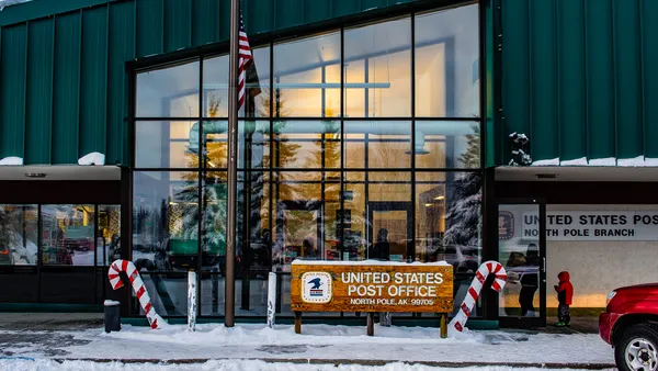 A United States Post Office sign sits in front of a building, as people enter the building.