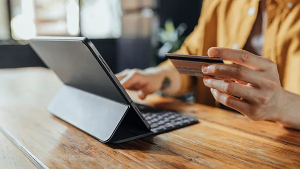 A seated person holds a payment card up as they type into a laptop placed on wooden table.