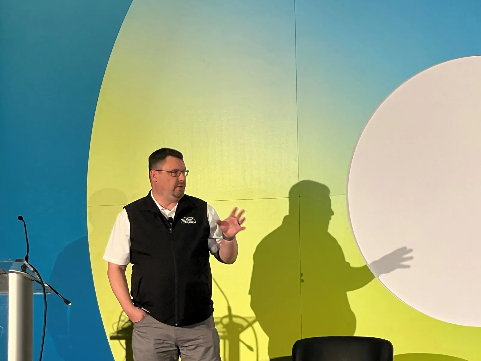 A photo of a person on stage in front of a white, yellow and blue wall speaking to an audience.