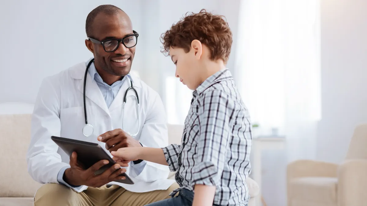 Male doctor smiling while showing tablet to boy and sitting.