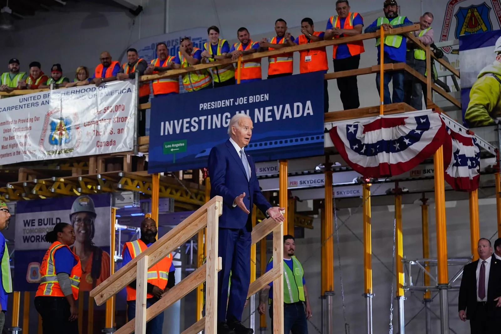 President Joe Biden climbing stairs to a podium surrounded by workers in construction vests with a sign reading &quot;Investing in Nevada.&quot;