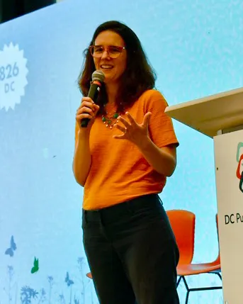 A person is standing a on a stage with a microphone. A large screen is in the background.