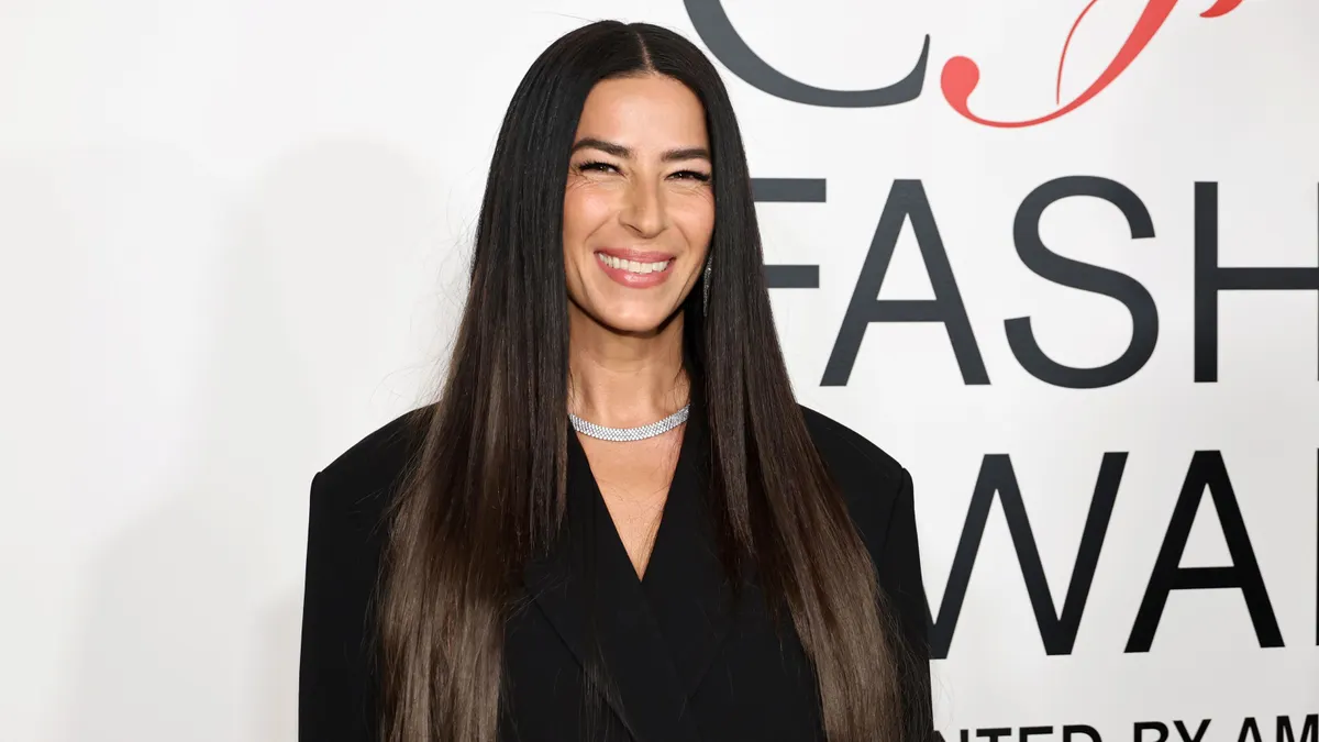 A person with long, straight, dark hair stands smiling at the camera in a close-up.