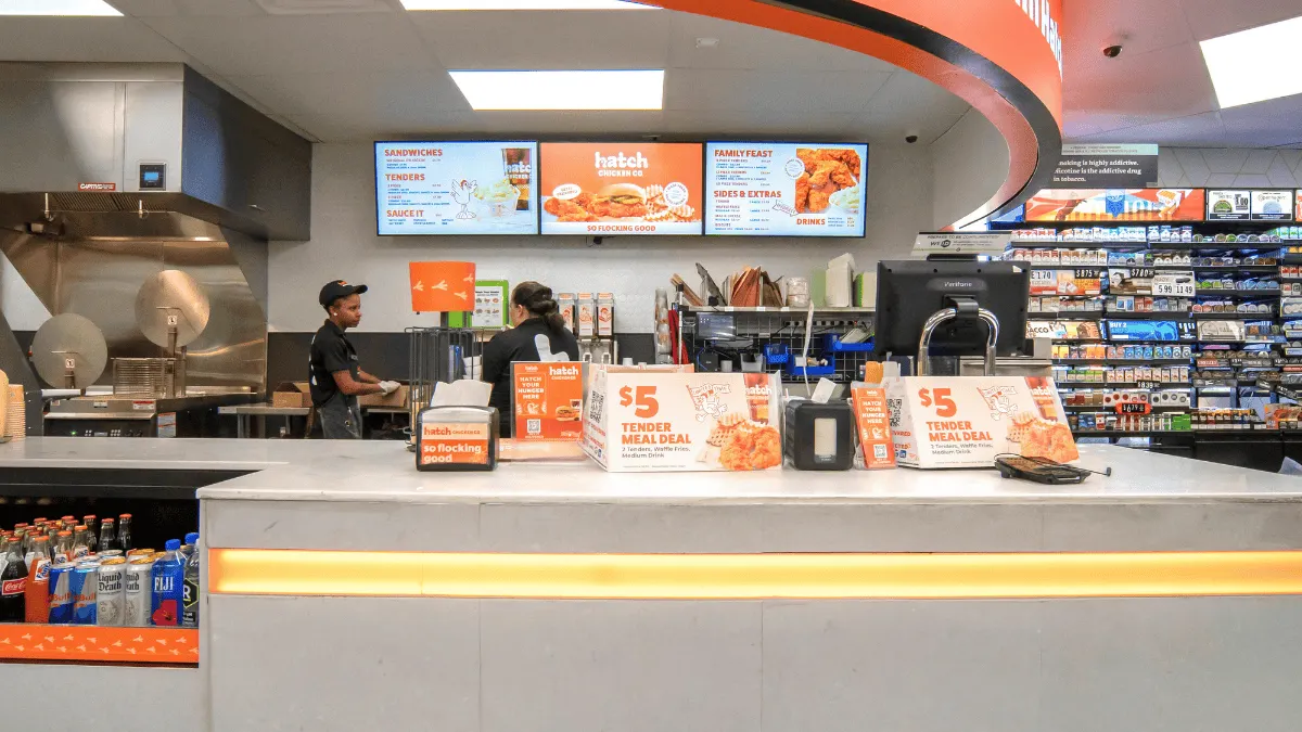 A photo of the interior of a convenience store, looking at a foodservice counter. A sign over the counter says "Brighten up your meals with Hatch."