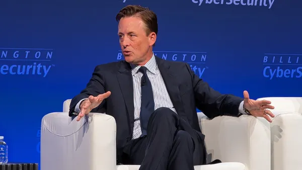 A man wearing a dark blue suit and tie gestures while sitting in a plush white chair in front of a backdrop that reads "Billington Cybersecurity"
