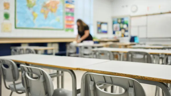 Chairs and desks sit in rows in a school classroom as a teacher works in the background.