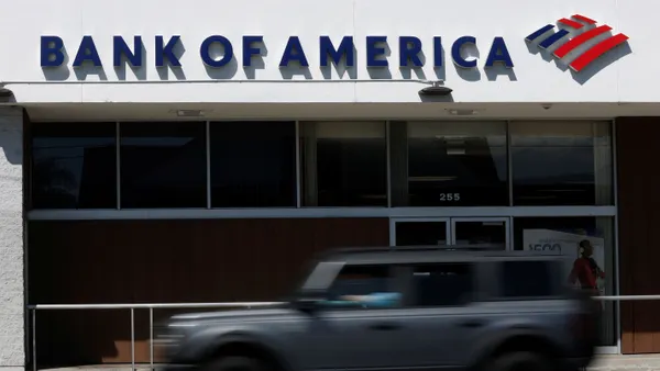 A car drives by a brick and glass storefront with the words "Bank of America" and the Bank of America flag logo above the entrance.
