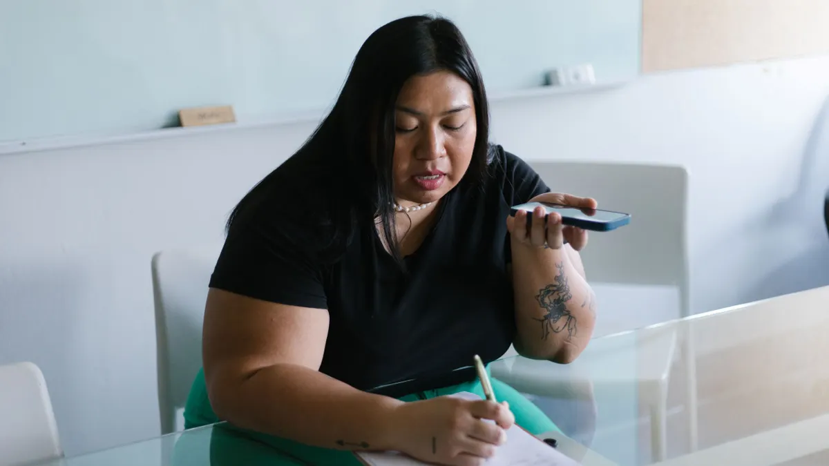 A person writes on papers on a clipboard while using speakerphone.