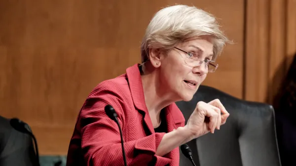 Sen. Elizabeth Warren, wearing a red jacket, speaks into a microphone during a Congressional hearing.