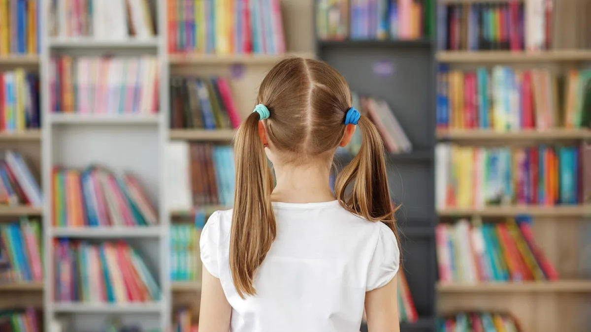 a young student with ponytails has their back to the camera and is facing a wall of shelves with multicolor books